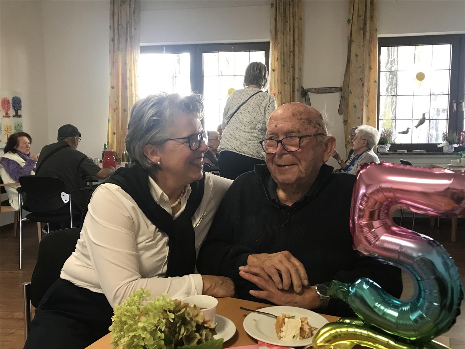Einrichtungsleitung der Tagespflege zusammen mit einem Gast und einem Luftballon als Zahl fünf