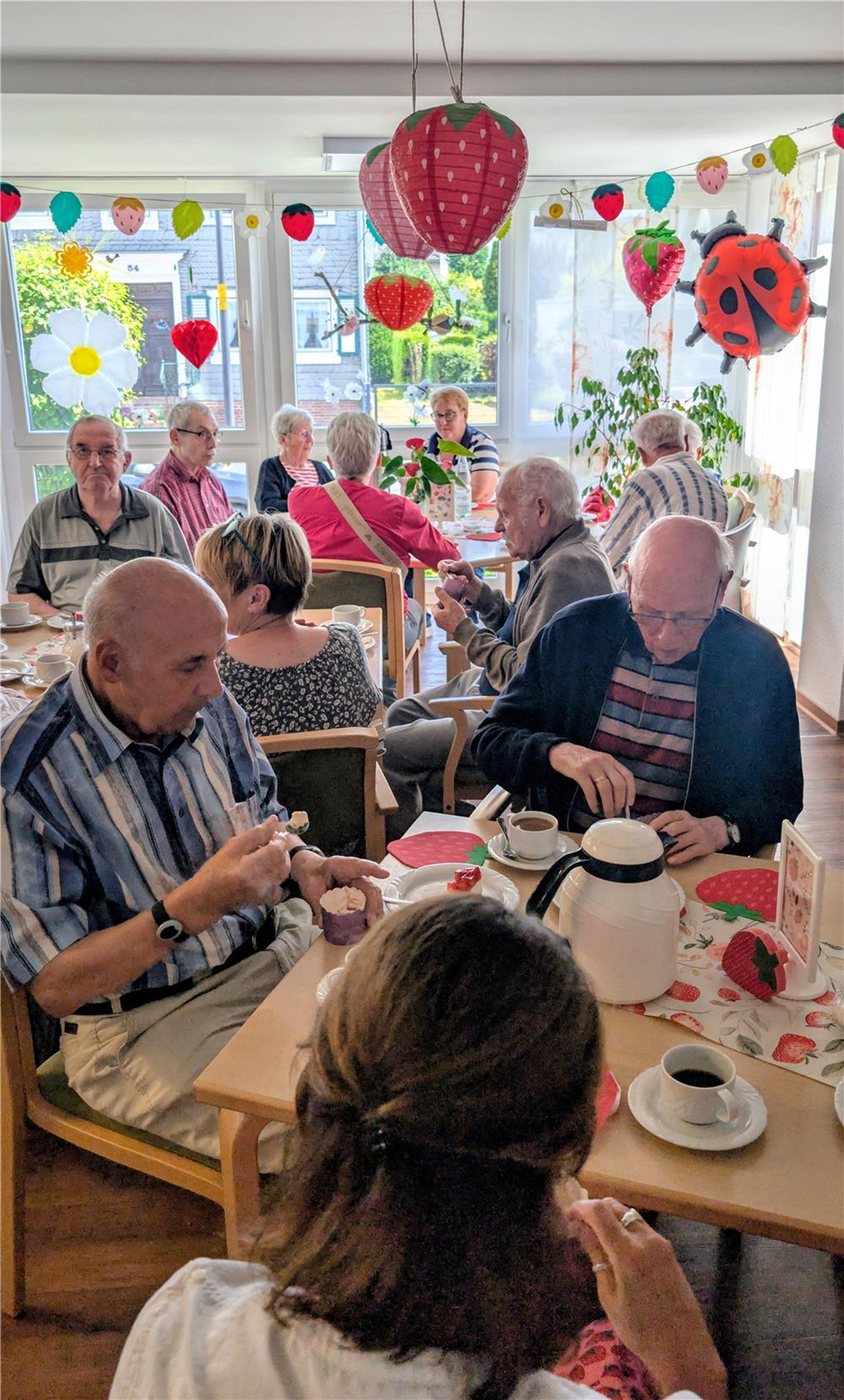 Gäste des Erdbeerfests sitzen bei Kaffee und Erdbeerdesserts zusammen. Erdbeerdekorationen schmücken den hellen Aufenthaltsraum und sorgen für eine sommerliche Stimmung.