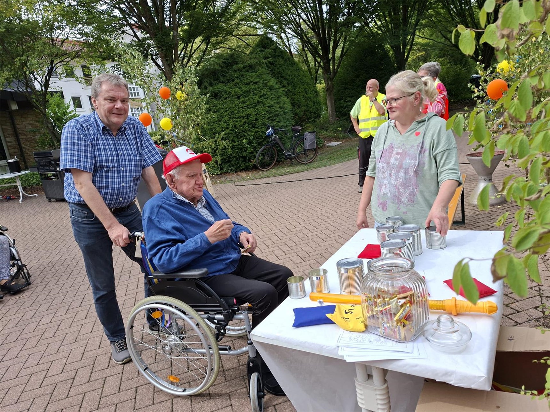Ein Bewohner nimmt gemeinsam mit einem Angehörigen am Dosenwerfen teil. Eine Mitarbeiterin betreut die Spielstation beim Baustellenfest im Innenhof des Seniorenhauses.