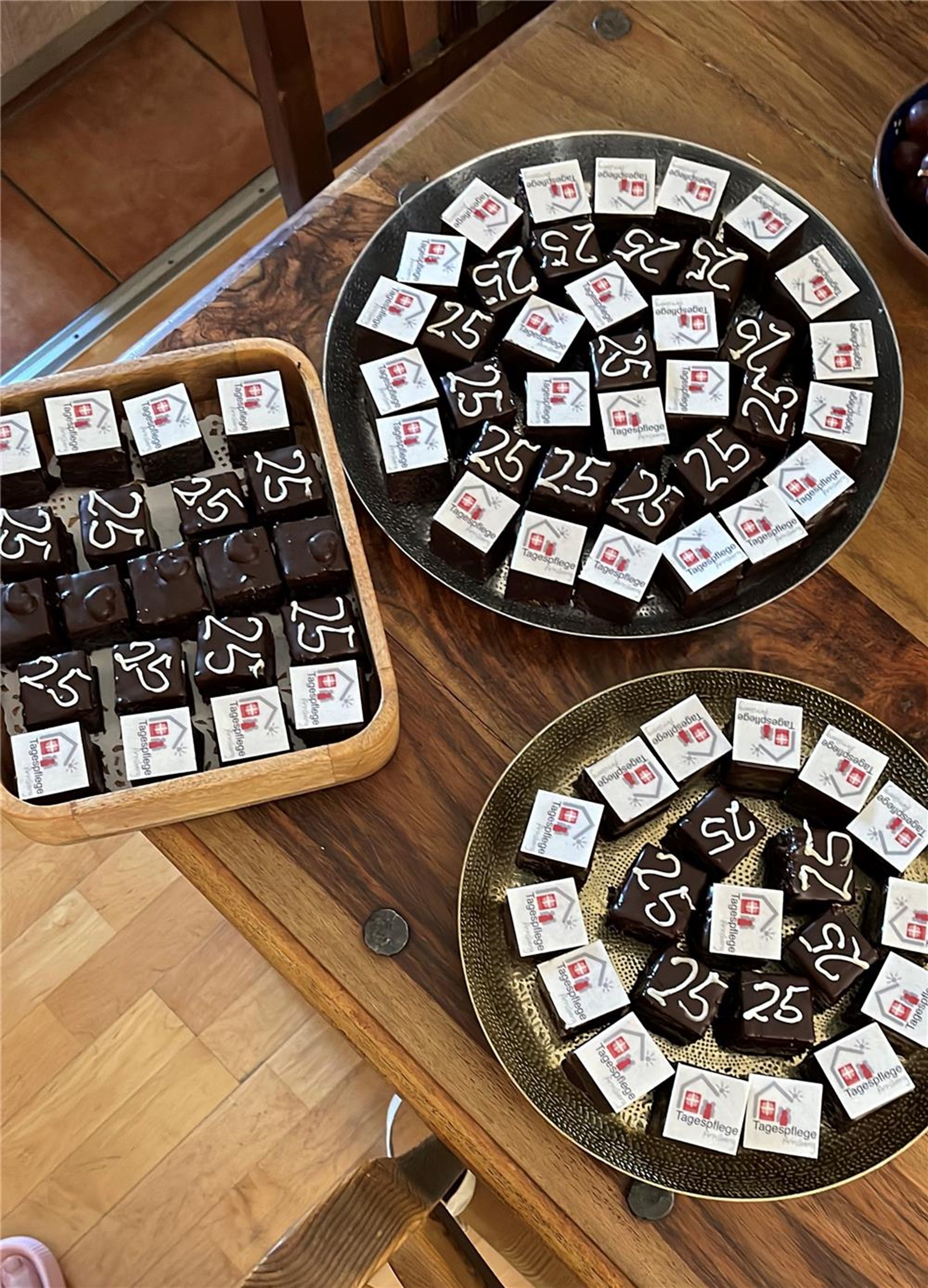 Kleine Schokoladentörtchen mit dem Logo der Tagespflege und der Zahl 25 sind dekorativ auf Platten angerichtet.