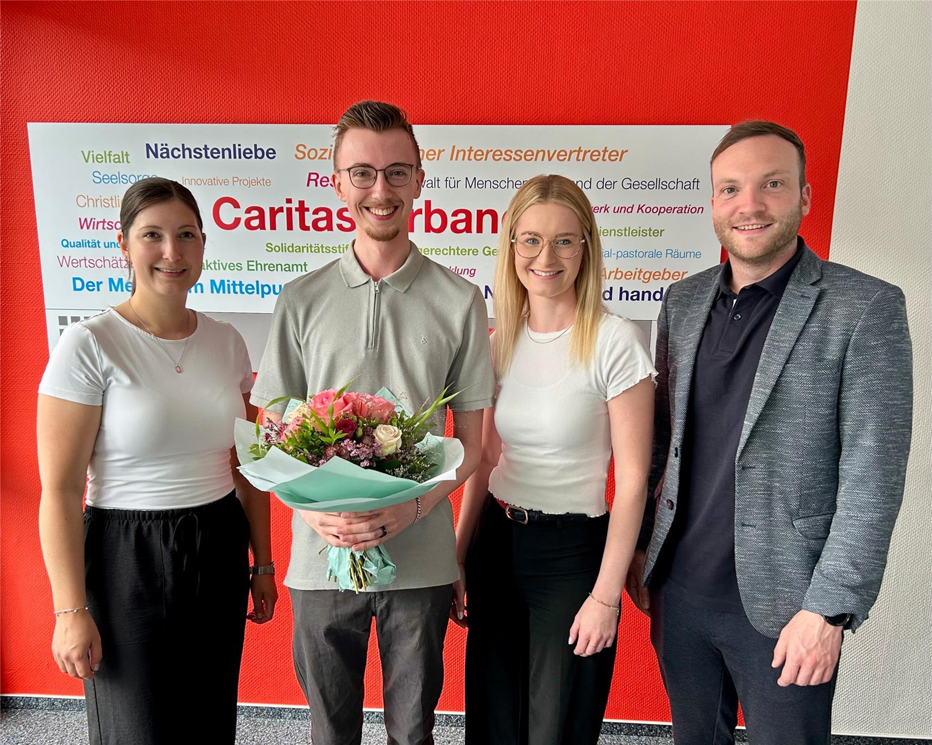 Vier Mitarbeitende des Caritasverbands stehen gemeinsam vor einer Caritas-Wand. Ein frisch ausgebildeter Kaufmann für Büromanagement hält einen Blumenstrauß in den Händen und wird von drei Kolleginnen und Kollegen zu seinem erfolgreichen Ausbildungsabschluss beglückwünscht.