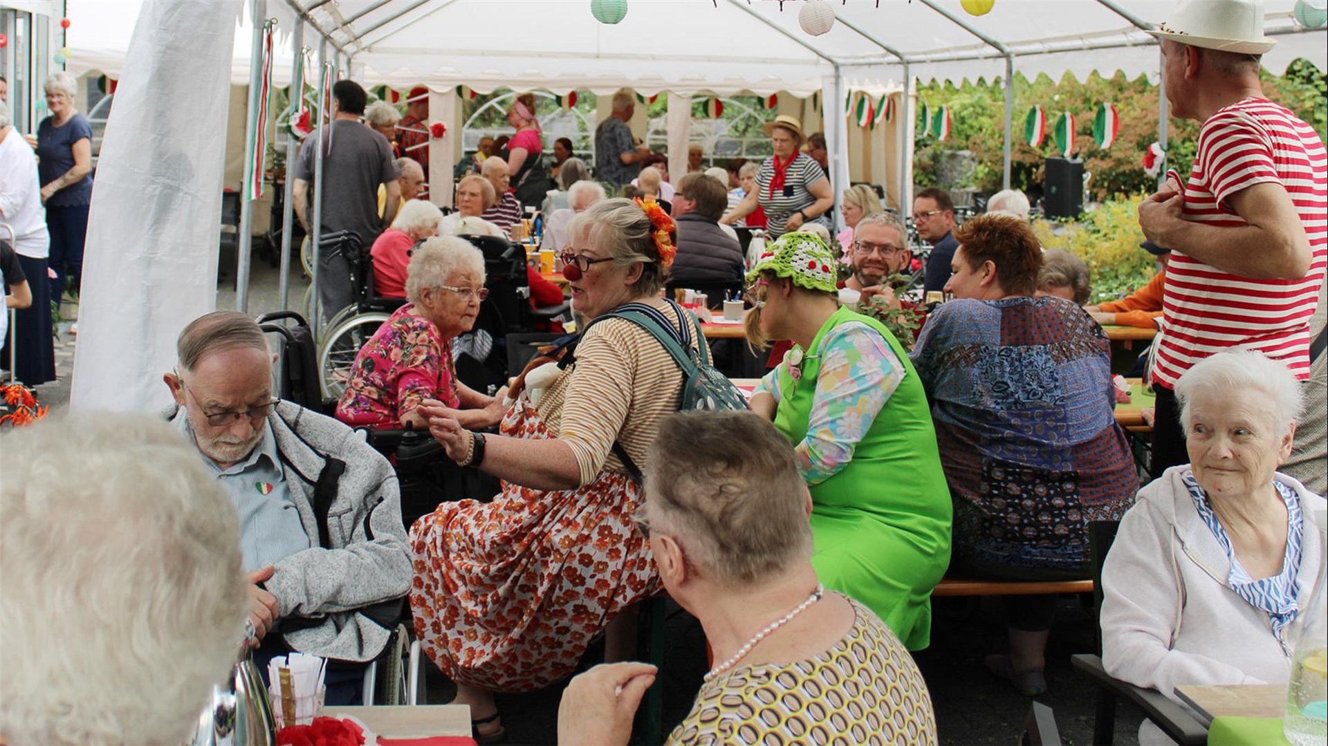 Zahlreiche Besucher und Besucherinnen, die Senior:innen des Seniorenhauses und traditionell italienisch gekleidete Personen sitzen unter einem Pavillon, der mit Wimpeln in den italienischen Nationalfarben geschmückt ist. 