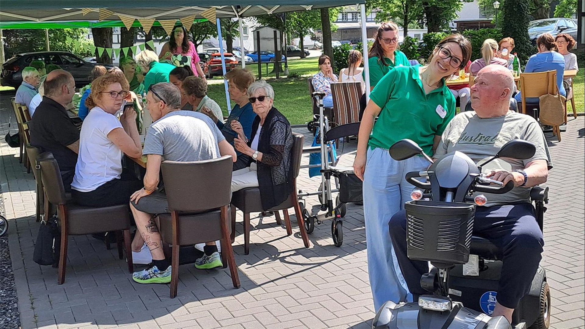 im Hintergrund sitzen Senior:innen und Betreuer:innen auf einer Terrasse bei Sonnenschein. Im Vordergrund ist ein Mann mit einem Scooter, eine Betreuuerin steht neben ihm und lacht in die Kamera. 