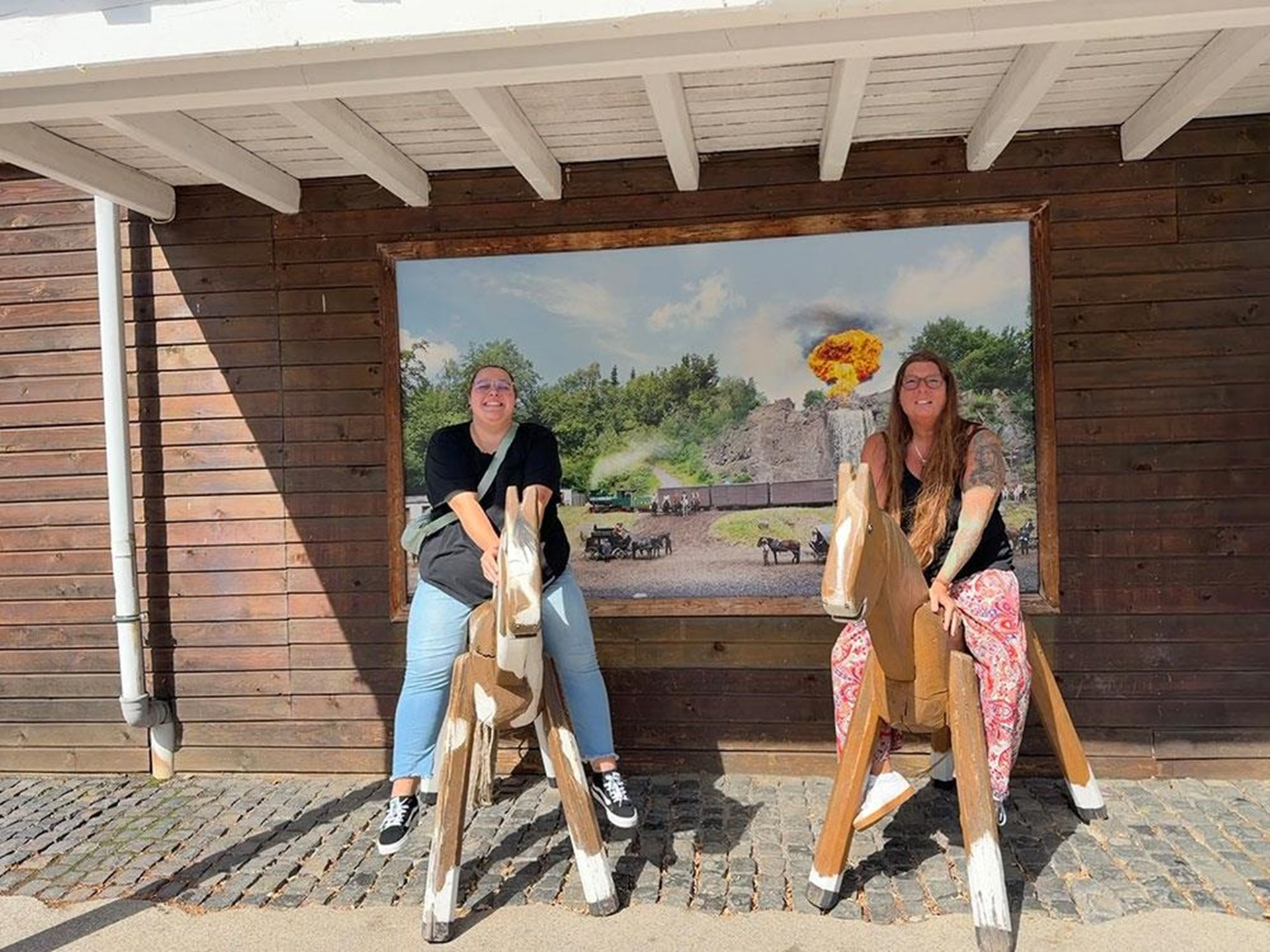 Zwei Mitarbeiterinnen sitzen lachend auf Holzpferden vor einer Westernkulisse.