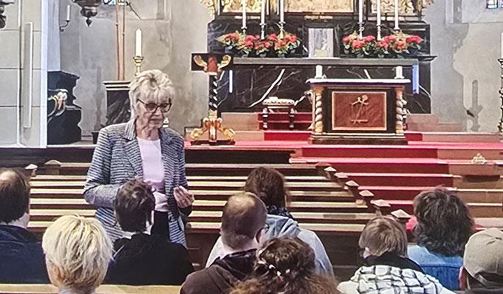 Beschäftigte der Caritas-Werkstätten Arnsberg und der EnsTeC sitzen während einer Führung in den Kirchenbänken der Propsteikirche St. Laurentius. Vor der Gruppe steht eine Frau und erläutert die Geschichte und Besonderheiten der Kirche. Im Hintergrund sind der historische Altar, Heiligenfiguren und die hohen, farbig verglasten Kirchenfenster zu sehen.