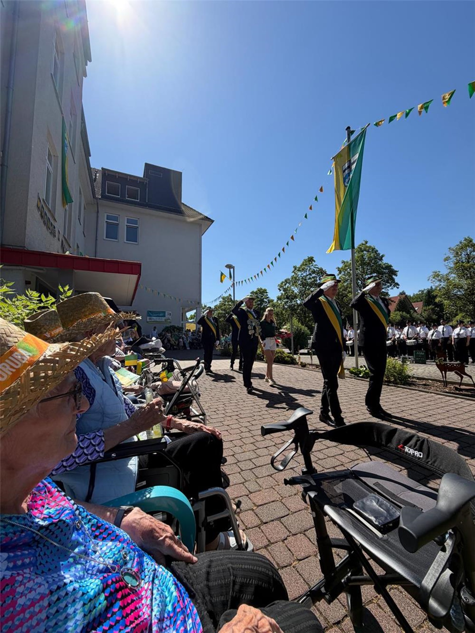 Bewohnerinnen und Bewohner sitzen in der ersten Reihe und verfolgen den Schützenfestumzug. Vor ihnen marschieren Schützen mit Fahnen und Musik durch die festlich geschmückte Straße.