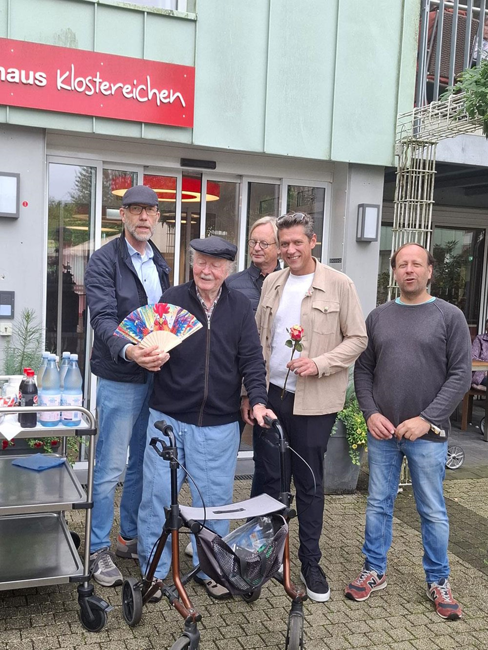 Mitglieder des Lions Club Neheim stehen gemeinsam mit einem Bewohner vor dem Eingang des Seniorenhauses Klostereichen. Sie halten einen bunten Fächer und eine Rose in den Händen.