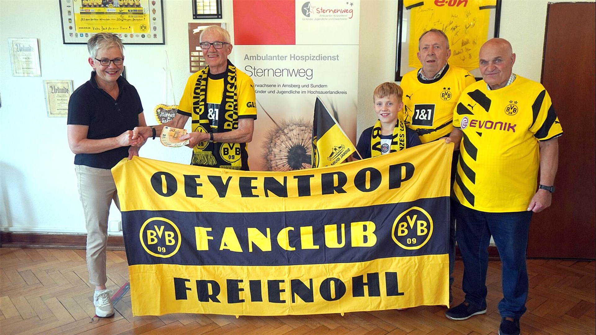 4 Erwachsene und ein Kind, Mitglieder des BVB Fanclubs Oeventrop und Freienohl halten ihr Vereinstransparent vor sich. Das Kind hält eine BVB-Fahne, einer der Erwachsenen übergibt den Betrag von 2000 Euro in Form von 50-Euro-Scheinen an die Mitarbeiterin des Sternenwegs. 