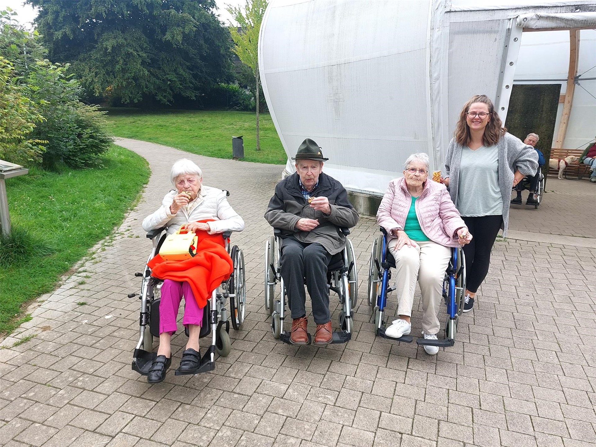 Senioren und Seniorinnen am Ufer eines Sees. Einige stehen, einige haben einen Rollator und einige sind im Rollstuhl. Auch Begleiterinnen stehen in der Gruppe. Alle lächeln. 