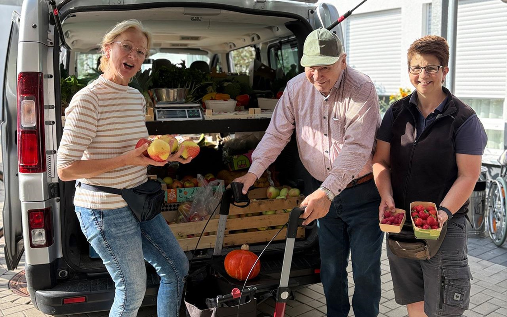 3 Personen stehen vor einem Verkaufswagen und halten frisches Obst und Gemüse in den Händen.